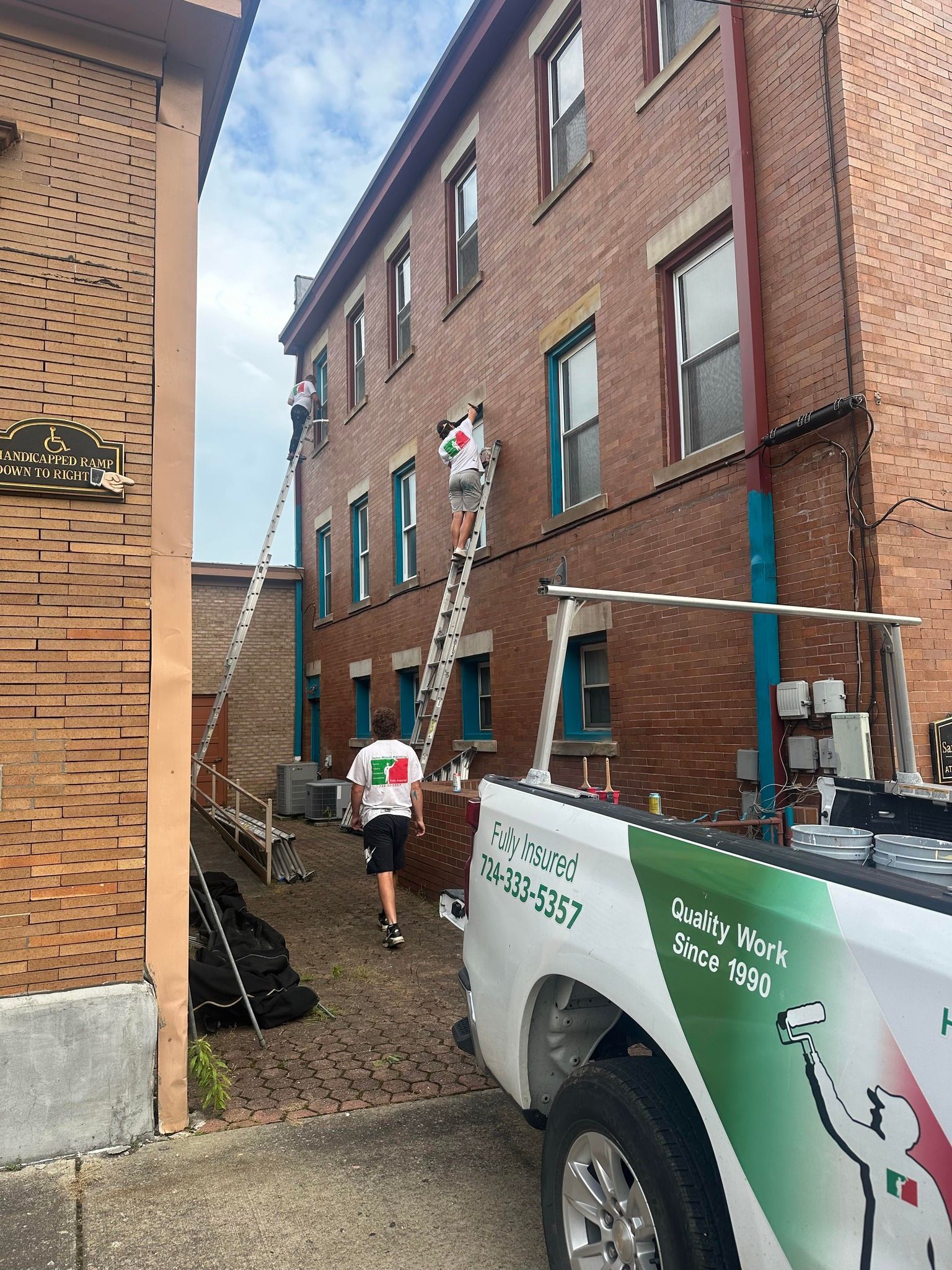 Two people on ladders working on a brick building with a truck parked nearby.