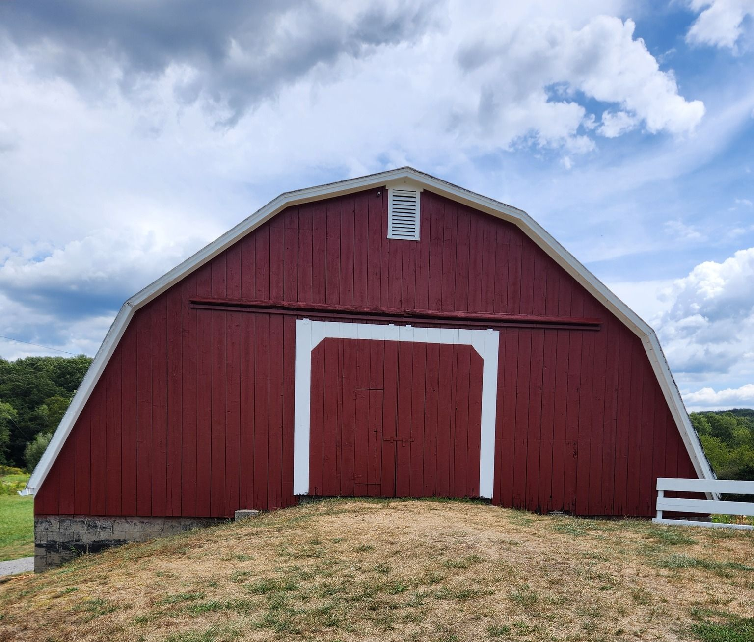 Red barn with white trim and a closed door, set on a small hill under a cloudy sky.