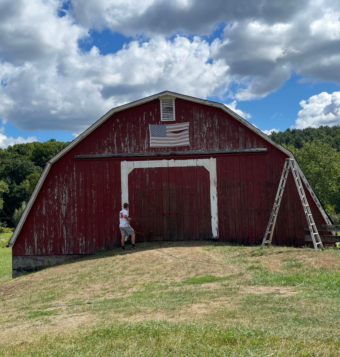 Red barn with white trim, a man standing in front of it, and a ladder on the side, under a cloudy blue sky.