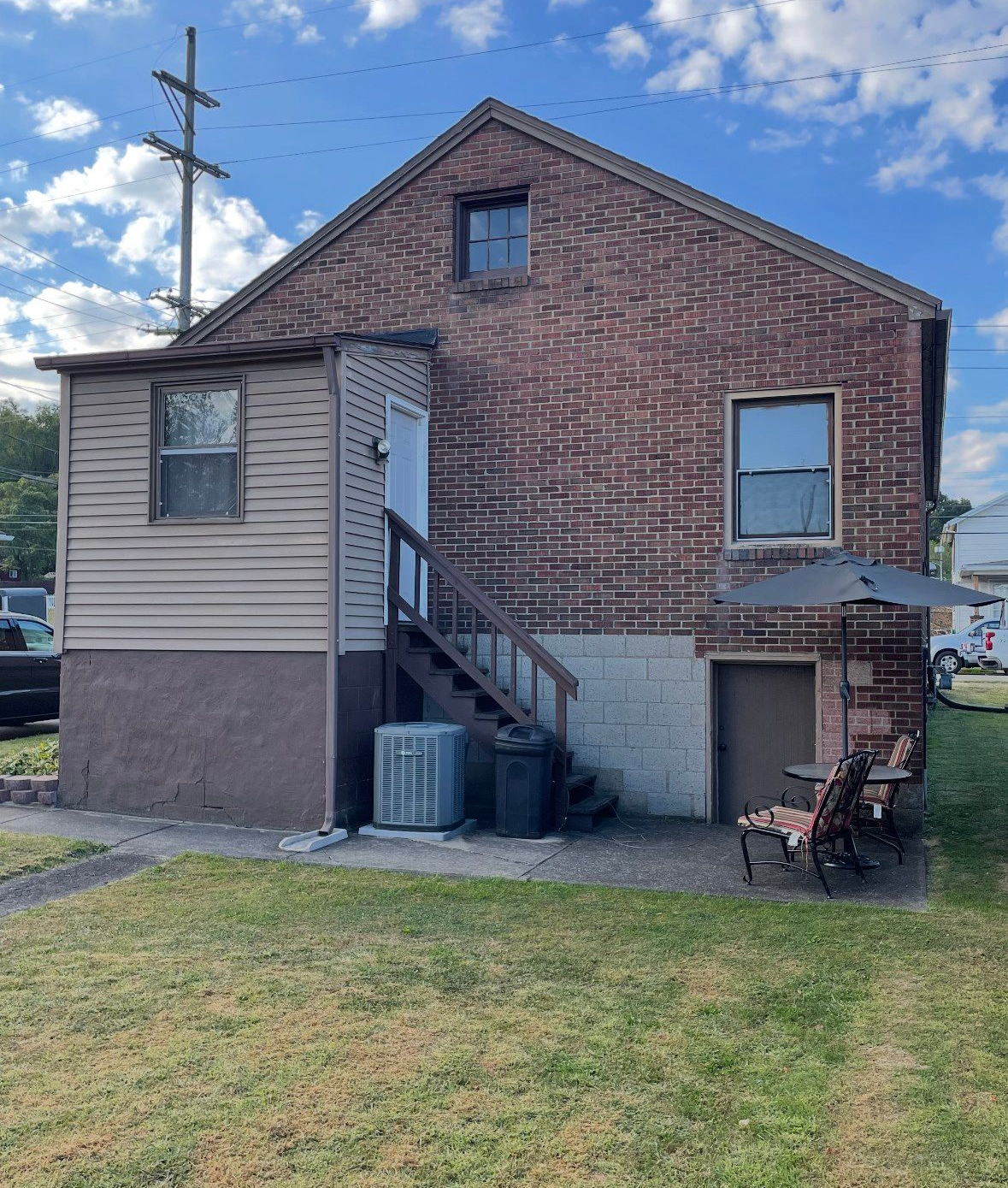 Back of a brick building with an attached porch, stairs, and yard. Air conditioner, door, windows, and chairs are visible.