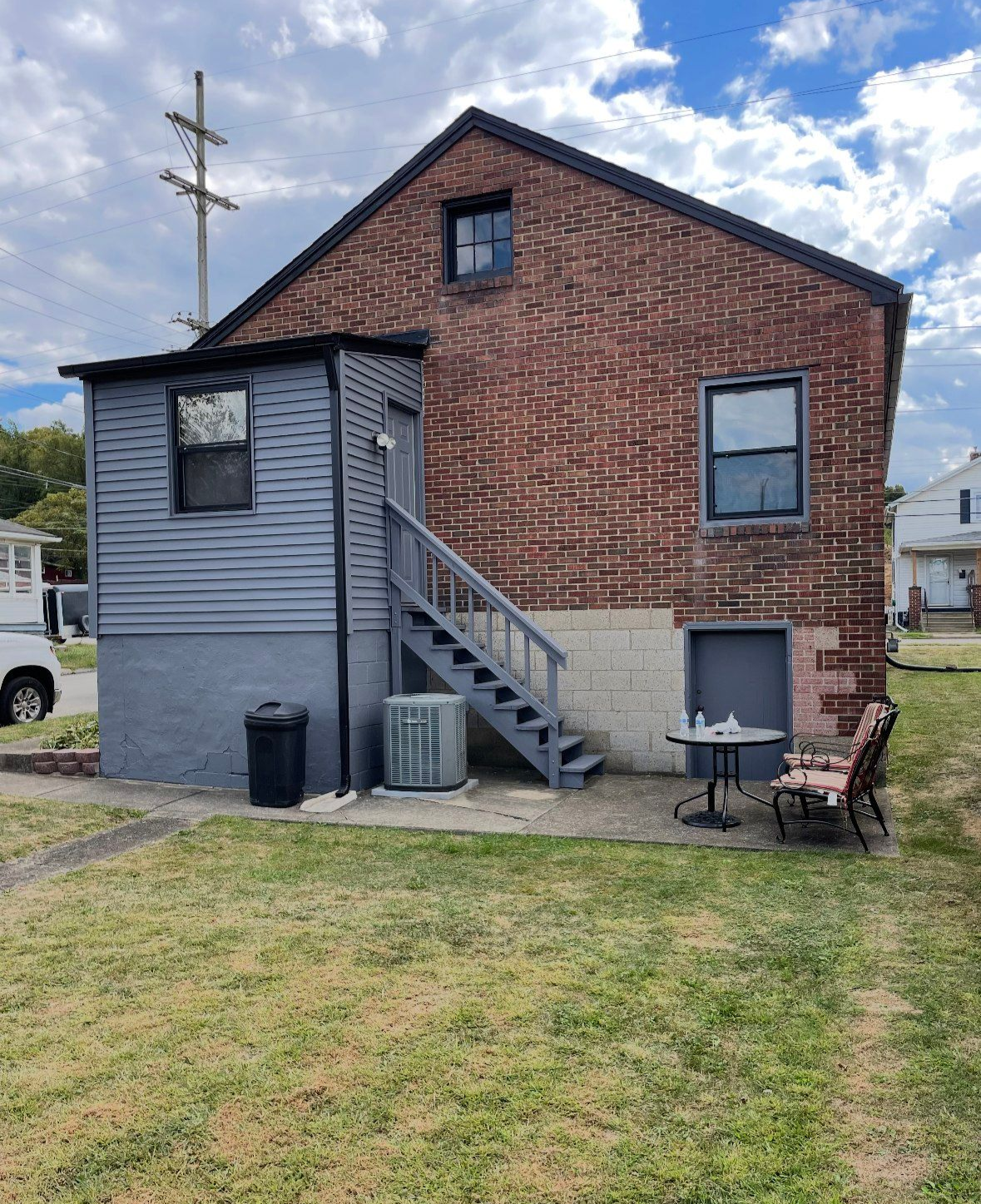 Back of a brick building with gray siding, stairs, and a small outdoor seating area.