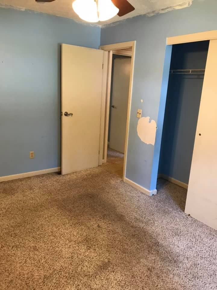 A light blue bedroom with tan carpet, a closet, open door to another room, and a damaged wall.
