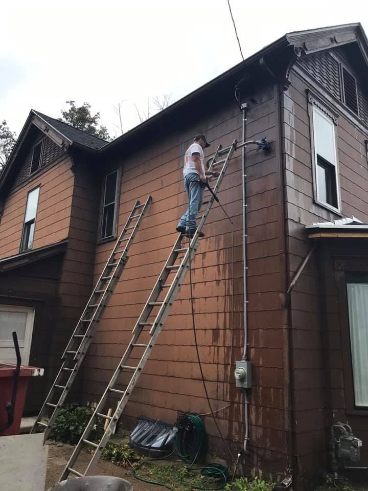 A person on a ladder washing the side of a brown house.