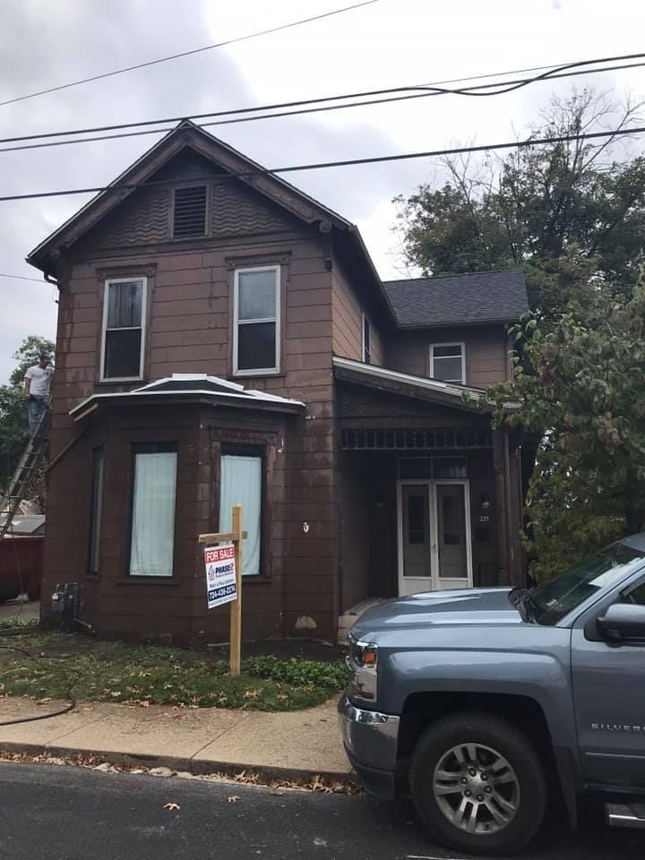 Brown two-story house with bay window and a person on a ladder; a sign and blue truck are in front.