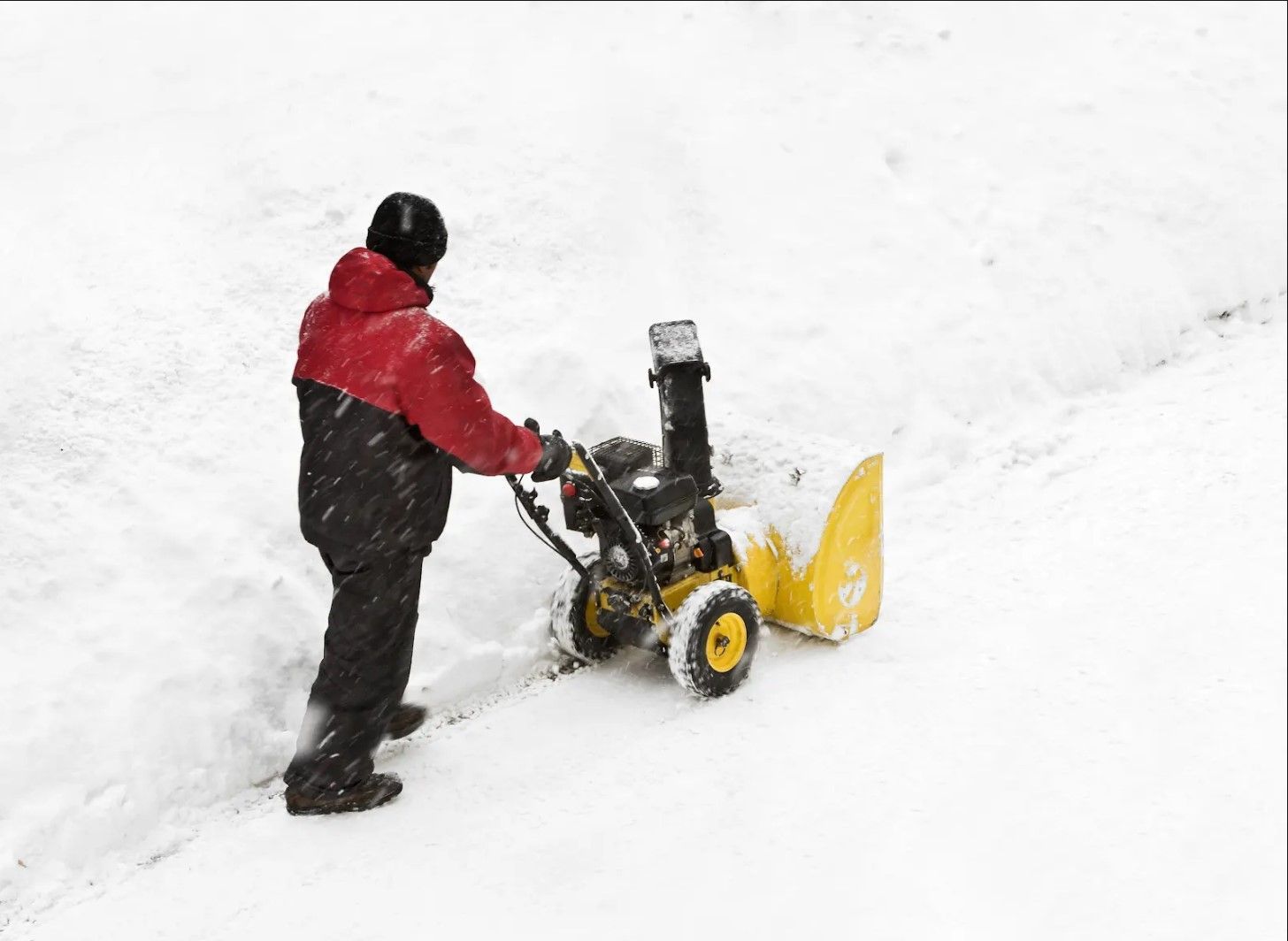Person in red jacket uses a yellow snowblower on a snow-covered surface, clearing a path.