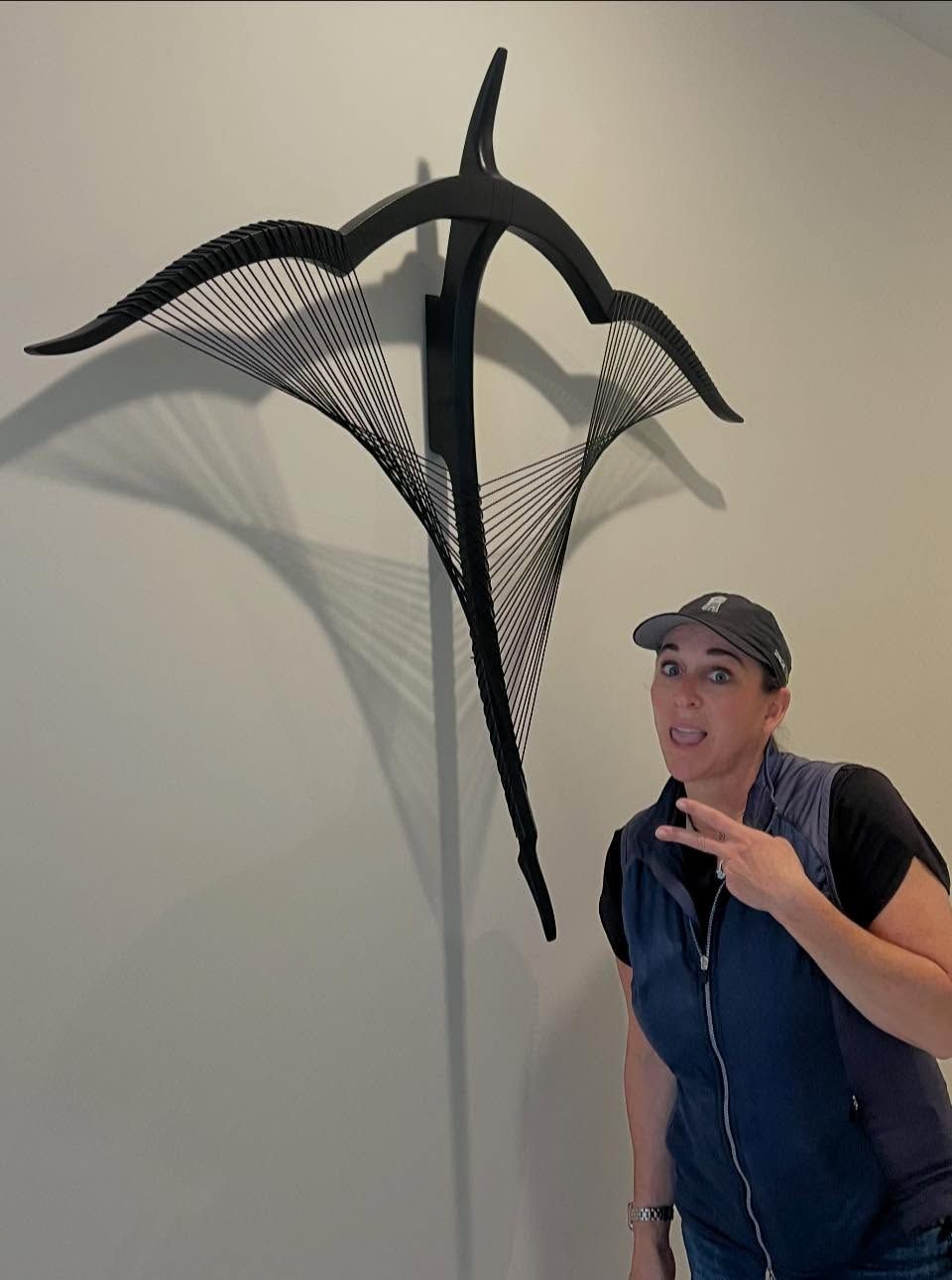 Woman posing beside a black abstract wall sculpture on a light-colored wall.