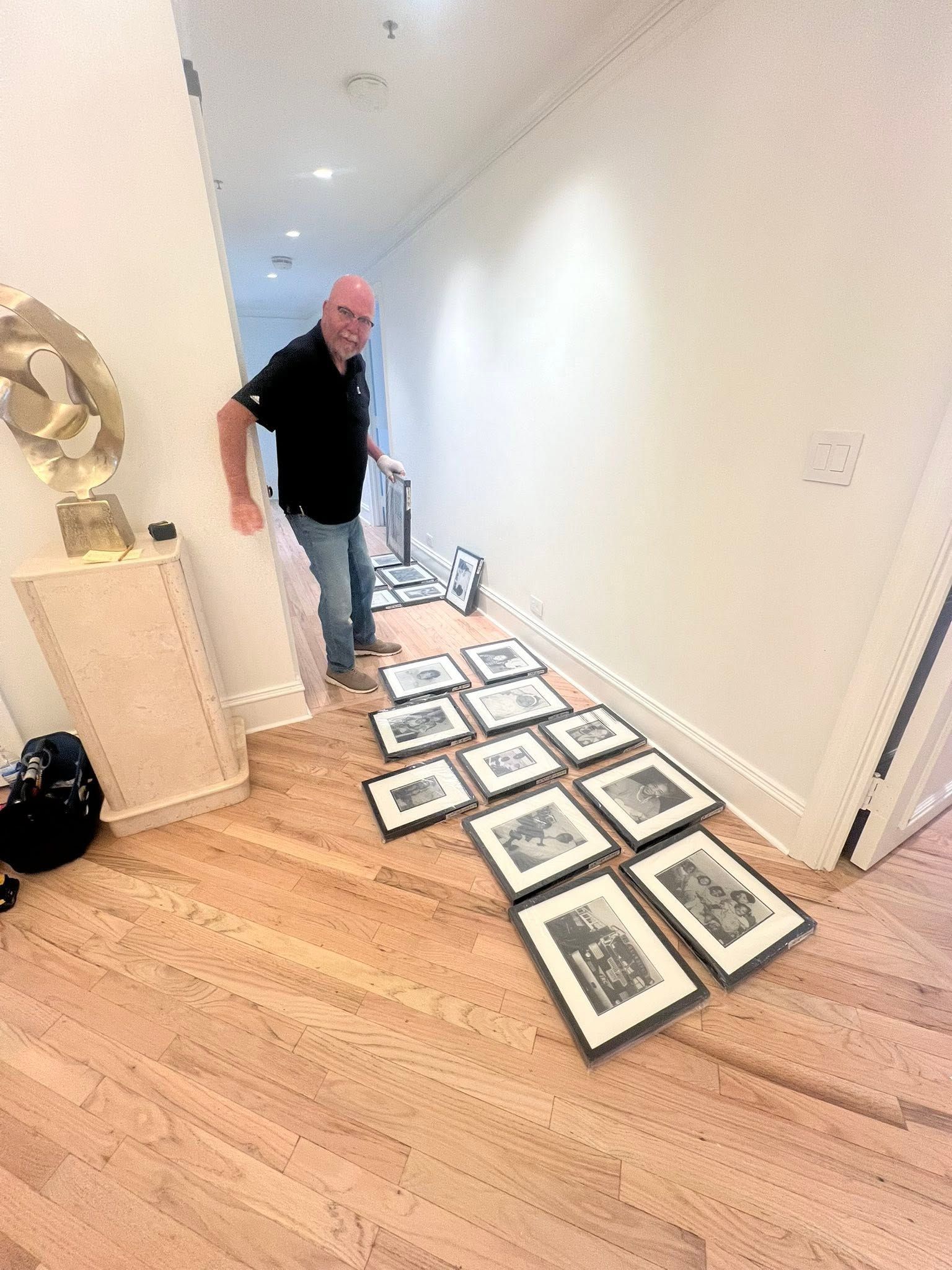 Man hanging framed photos in a hallway beside a wall of paintings laid on the floor