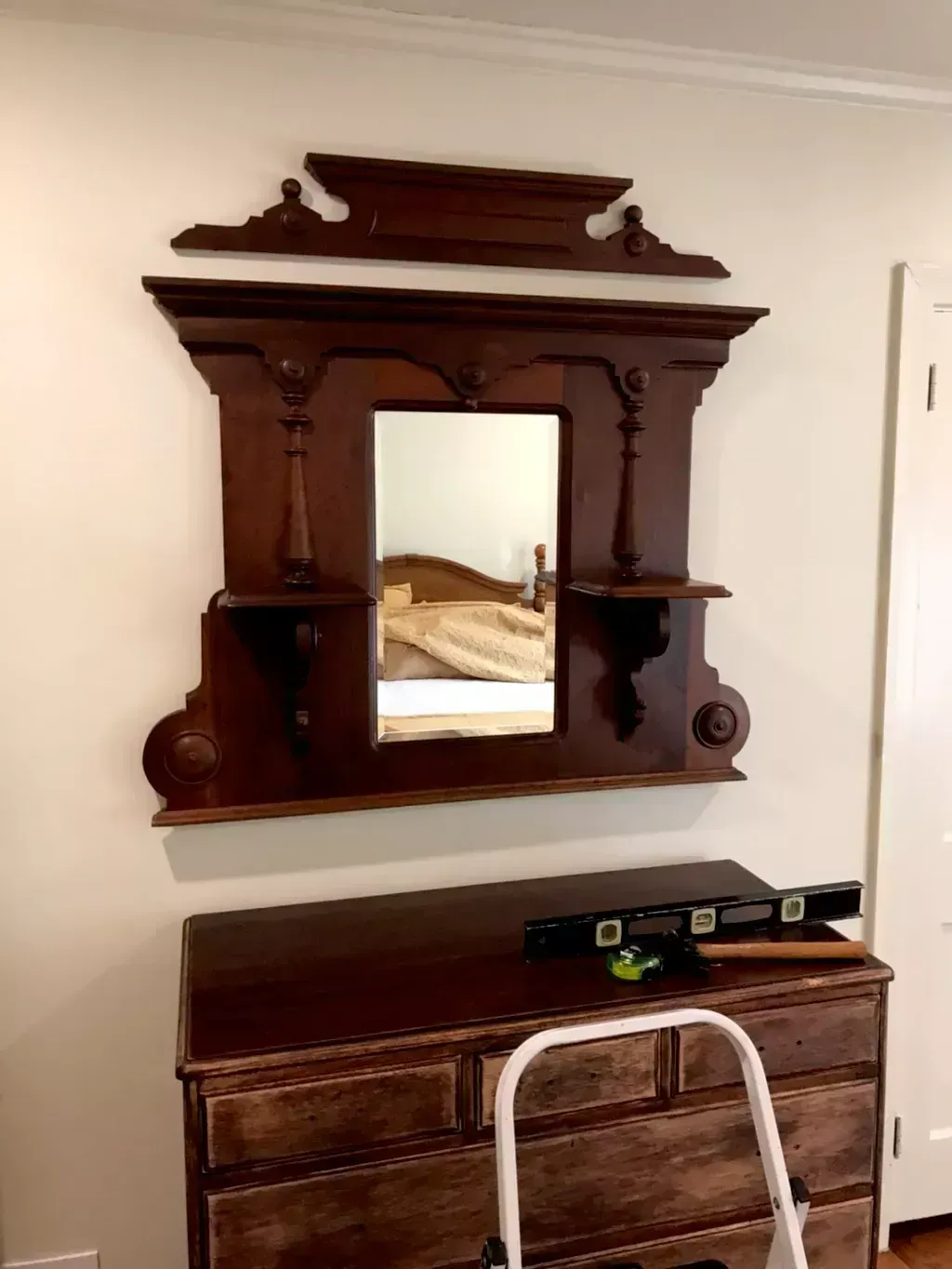 Antique dark wood wall mirror above a dresser in a beige room, with a step stool in front.