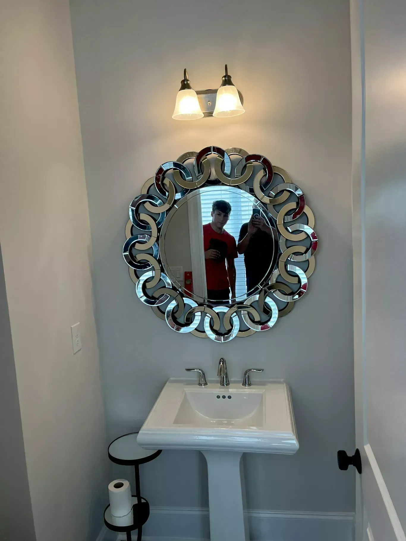 Small bathroom vanity with ornate round mirror, two-bulb light, sink, and a person reflected in the mirror