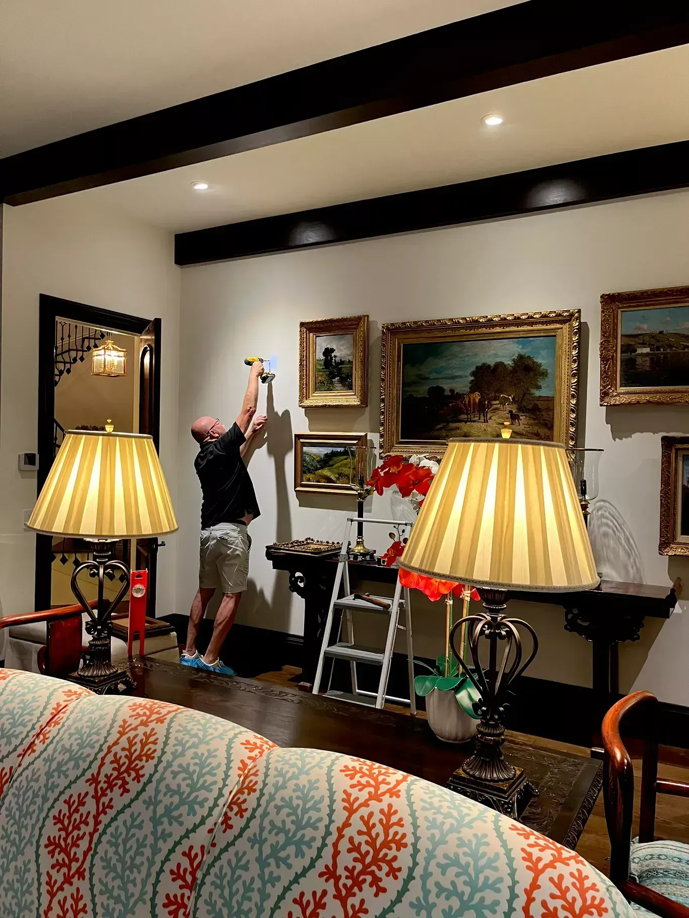 Person hanging framed art on a wall in a warm living room with lamps and flowers.