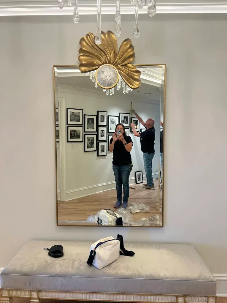 Mirror in a stylish room reflects two people, a gallery wall, and a gray bench with items on it.
