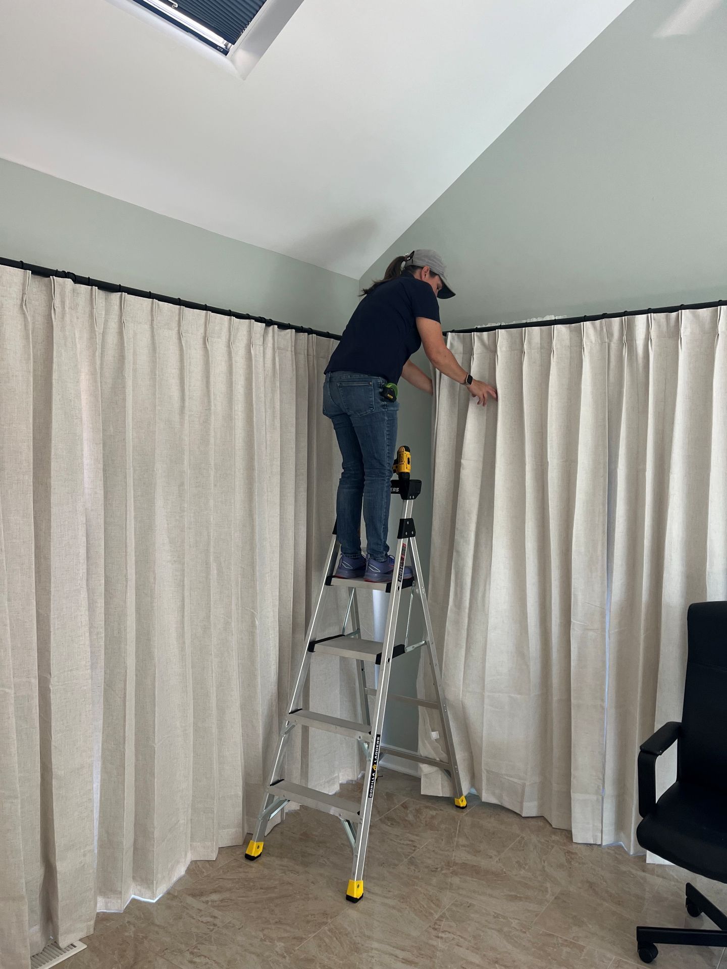 Person on a ladder adjusting curtains in a bright room.