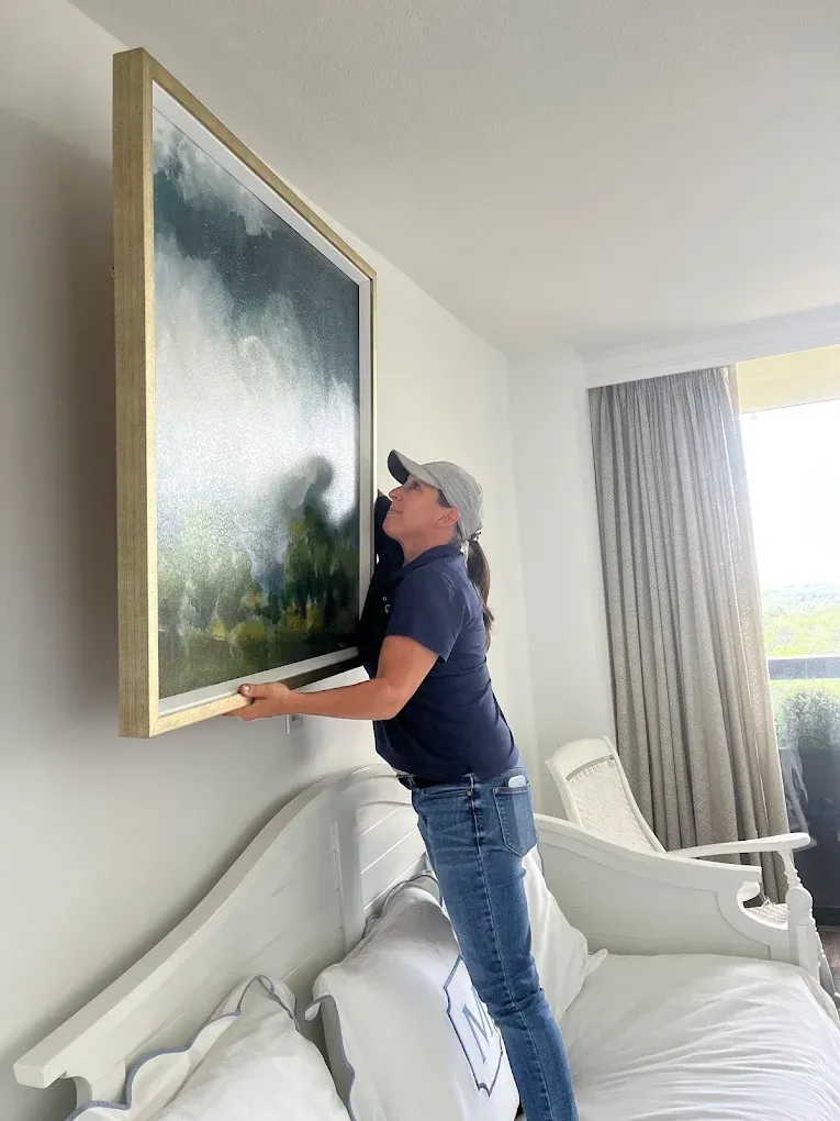 Person hanging a large framed painting on a wall beside a bed in a bright bedroom.
