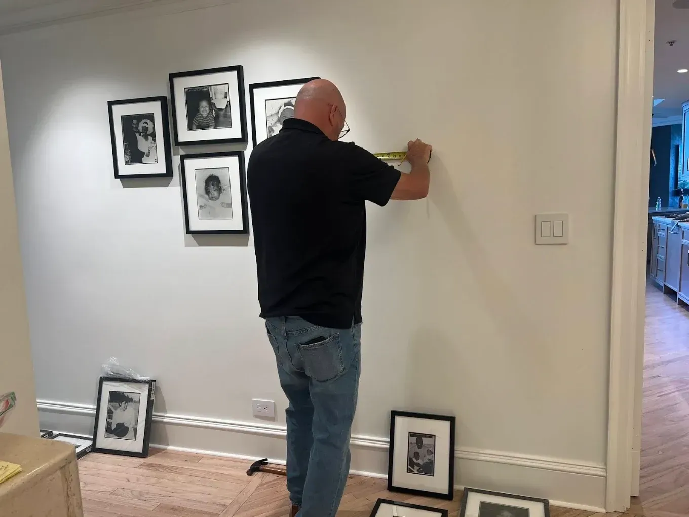 Man hanging framed photos on a wall in a hallway with more frames leaning below