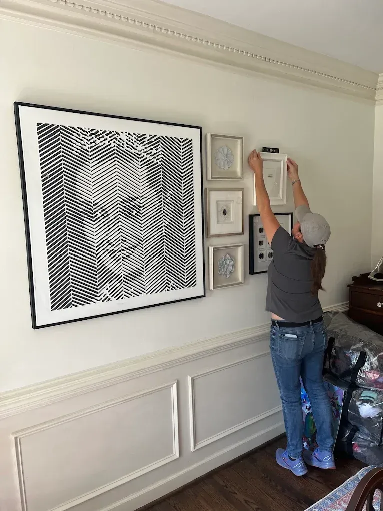 Person hanging framed art in a white-paneled room with a large portrait on the wall