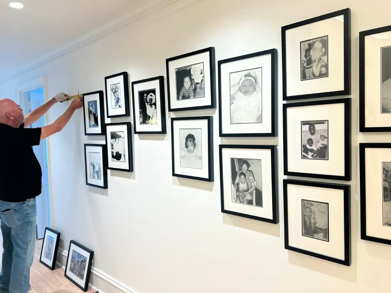 Person hanging framed black-and-white art in a white hallway gallery wall