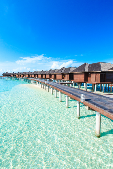 A wooden walkway leads to a long row of overwater bungalows in a bright blue, tropical ocean under a clear sky.