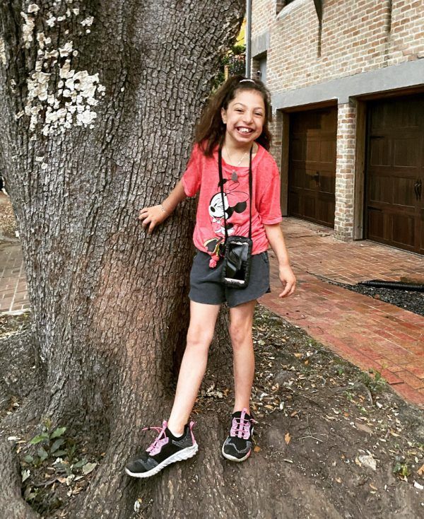 A young girl is standing next to a tree with a camera around her neck.