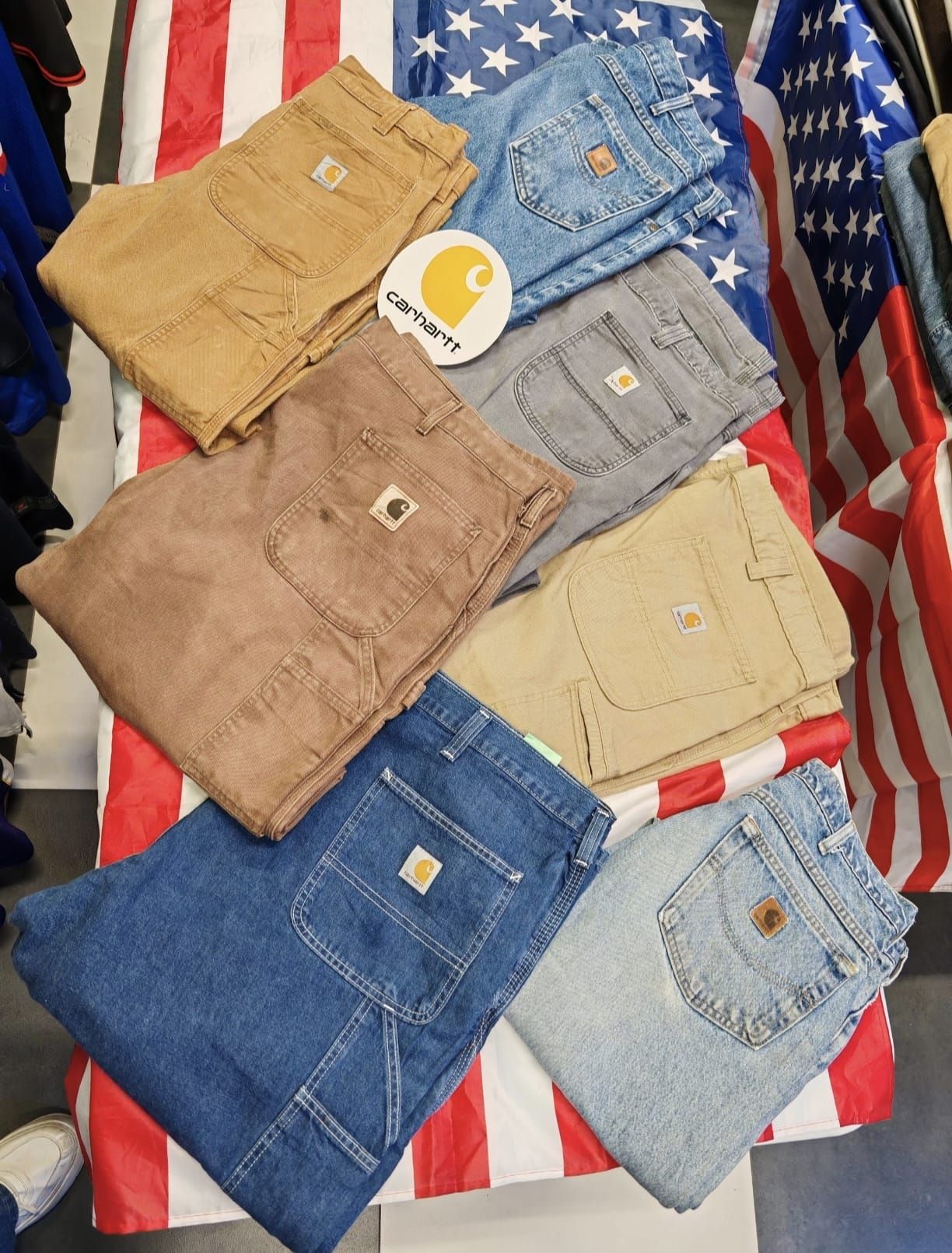 Vari modelli di jeans piegati nei colori beige, marrone, grigio, blu e azzurro su uno sfondo con la bandiera americana rossa, bianca e blu.
