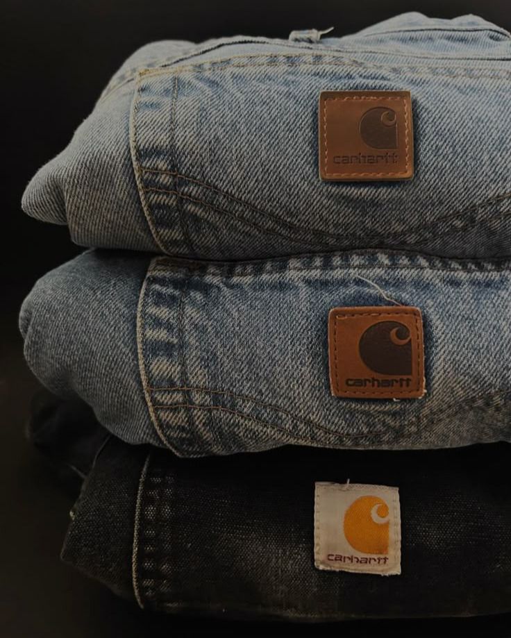 Tre paia di jeans sovrapposti, in lavaggio chiaro, medio e scuro, con toppe Carhartt.