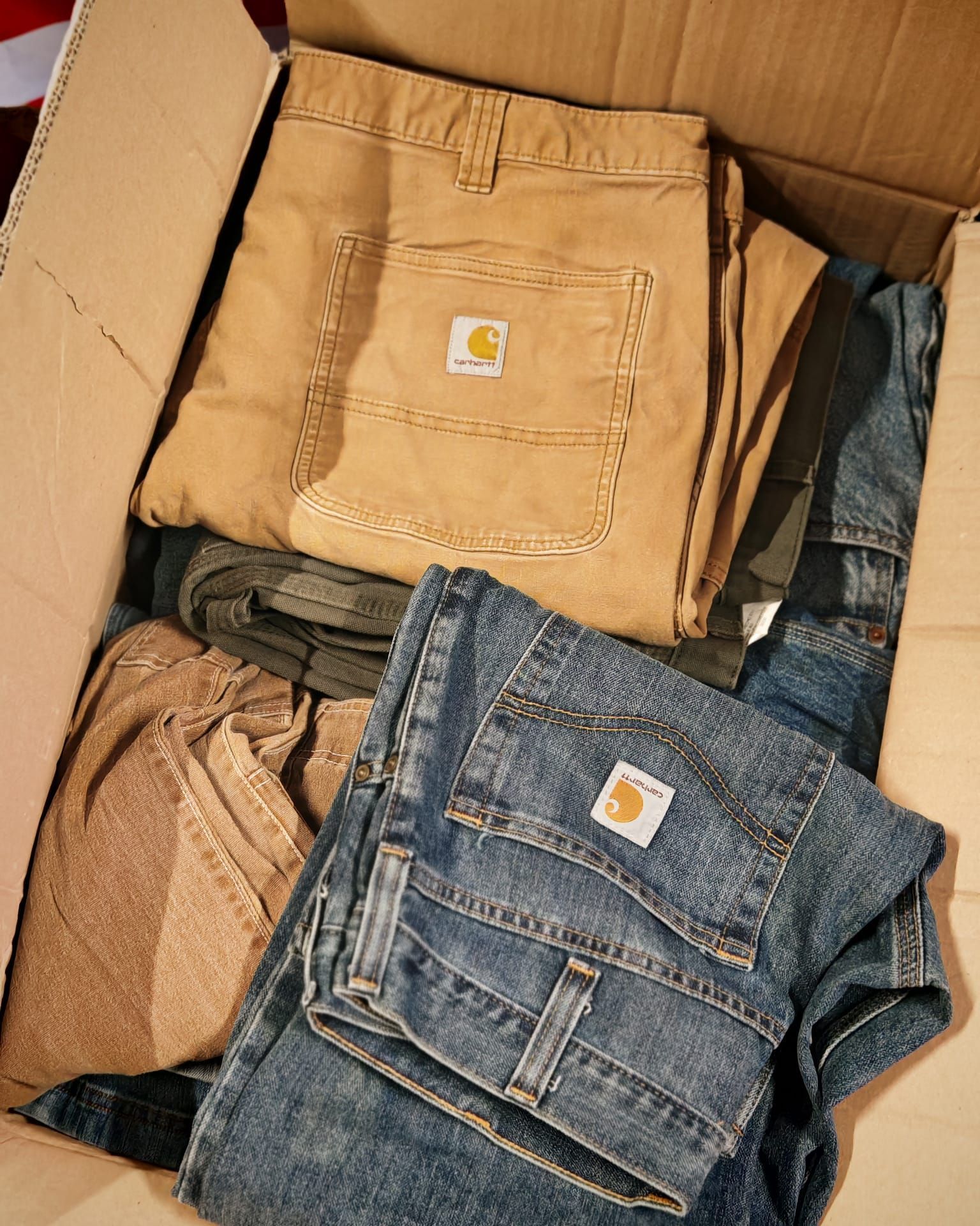 Scatola di cartone aperta piena di pantaloni e jeans piegati in denim beige, nero e blu.