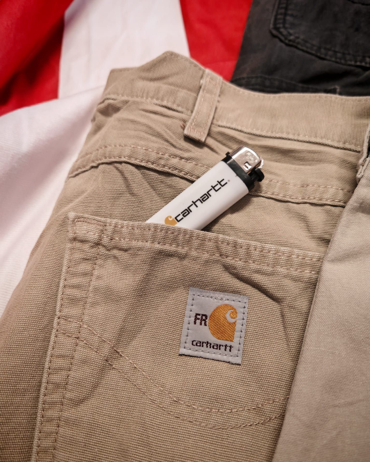 Tasca di pantaloni Carhartt beige con accendino, primo piano sul tessuto testurizzato e sulla toppa del logo