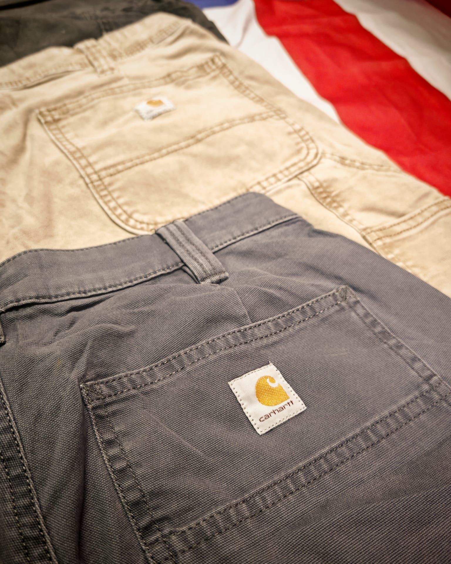 Due pantaloni da lavoro Carhartt sovrapposti, uno color cachi e uno grigio, con tasche posteriori e patch con logo ben visibili.