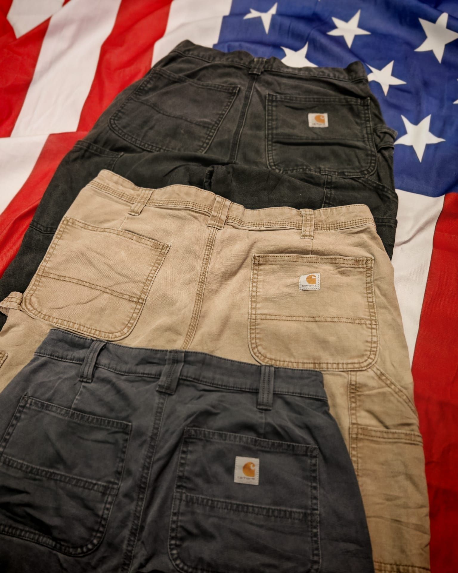 Tre paia di pantaloni Carhartt sovrapposti, nei colori nero, beige e grigio, su uno sfondo con la bandiera americana.
