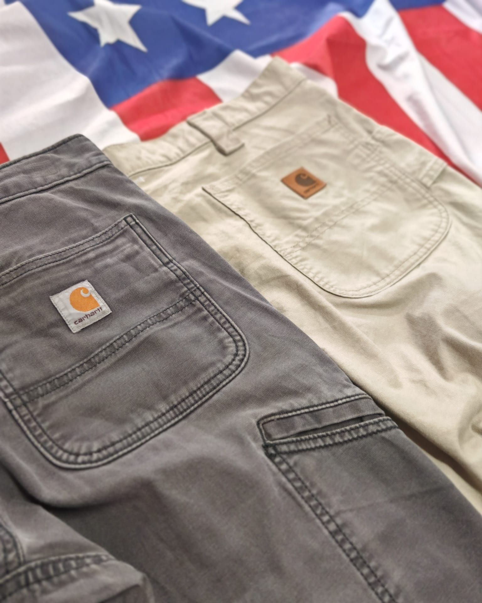 Tre paia di pantaloni Carhartt nei colori grigio, beige e nero su uno sfondo con la bandiera americana.