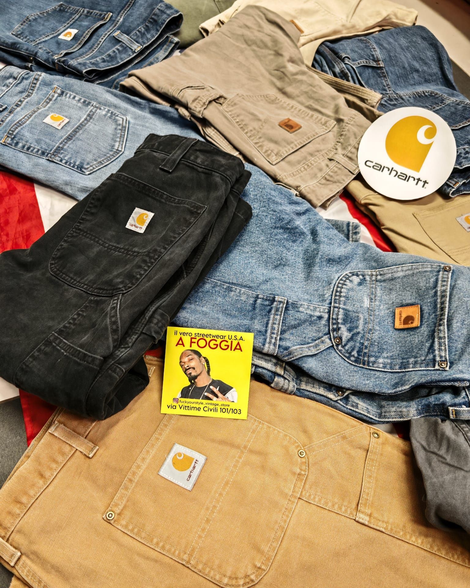 Assortiti jeans e pantaloni Carhartt nei colori blu, beige e nero, impilati uno sull'altro, con le toppe del logo ben visibili.