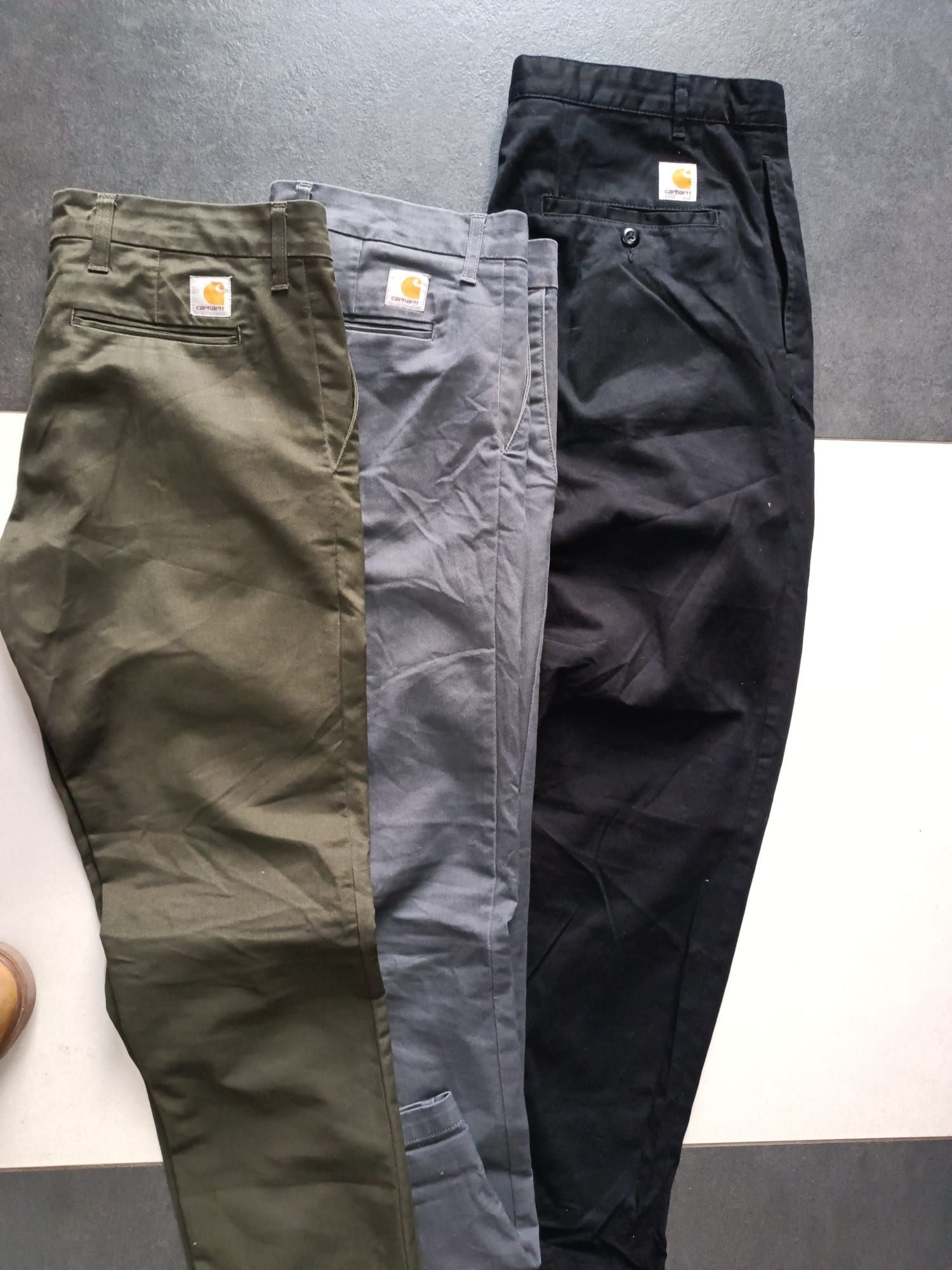 Tre paia di pantaloni cargo verde oliva, grigi e neri disposti uno accanto all'altro su una superficie bianca.