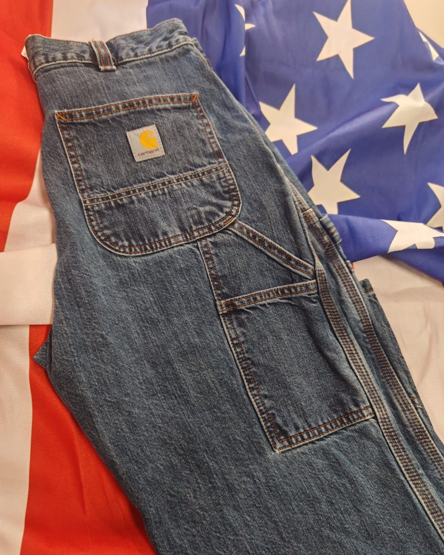 Jeans blu piegati con una toppa con logo giallo, adagiati su un tessuto con motivo a stelle rosse, bianche e blu.