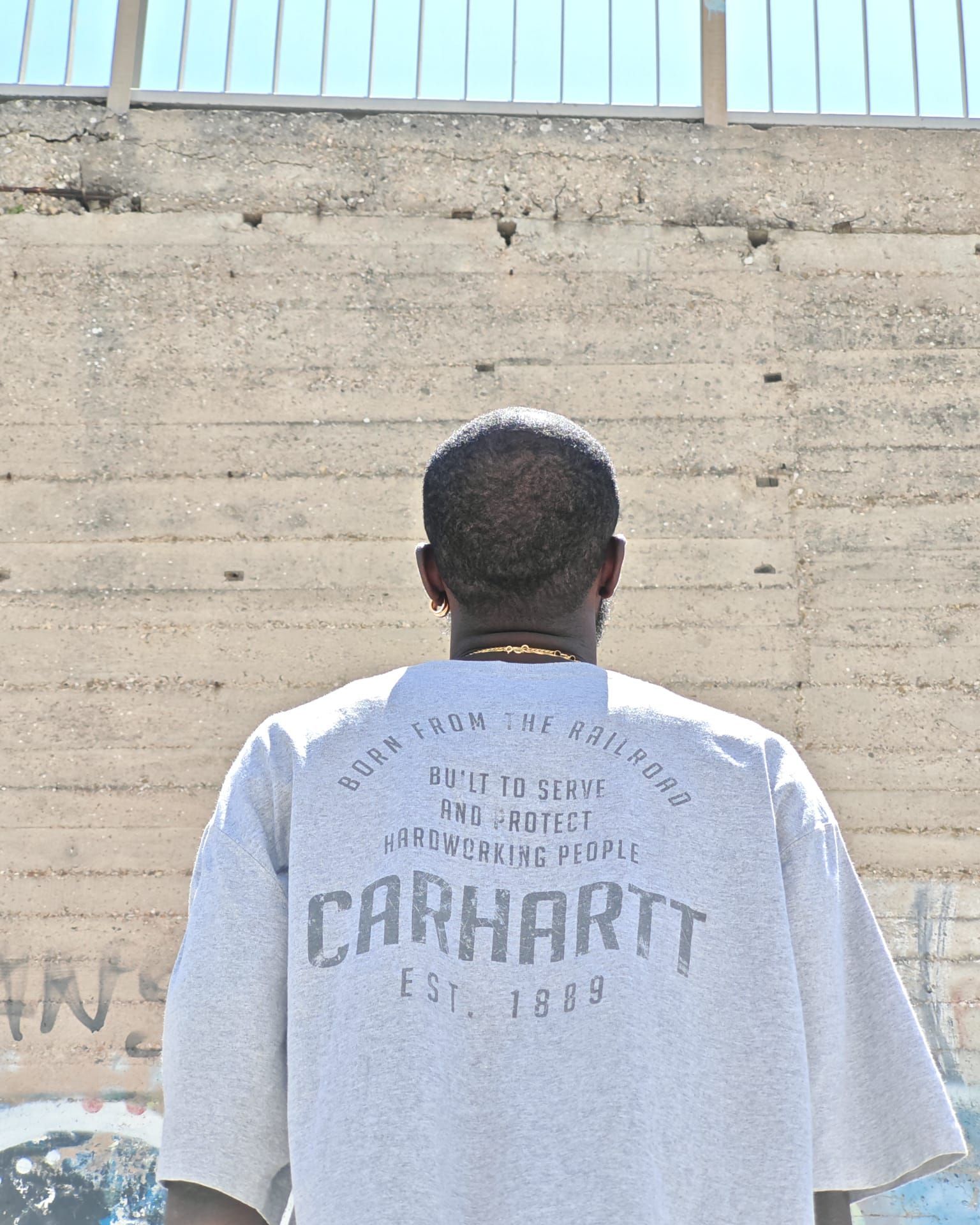 Persona vista di spalle, con indosso una maglietta grigia della Carhartt, in piedi contro un muro di cemento all'aperto.