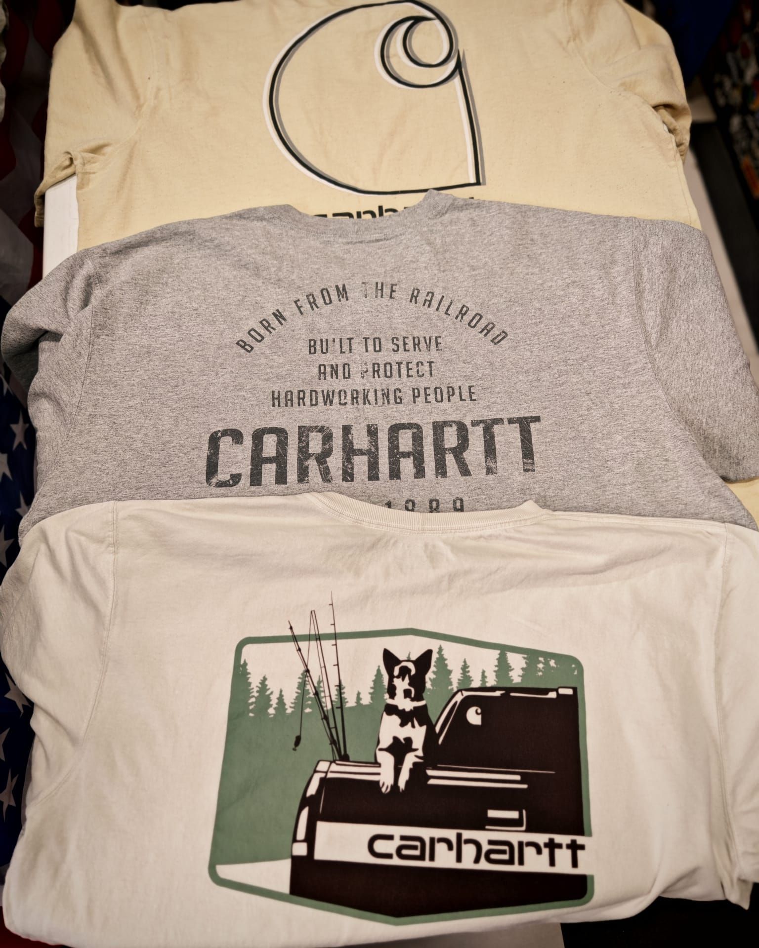 Tre magliette Carhartt piegate e impilate, beige e grigie, con loghi e la grafica di un cane.