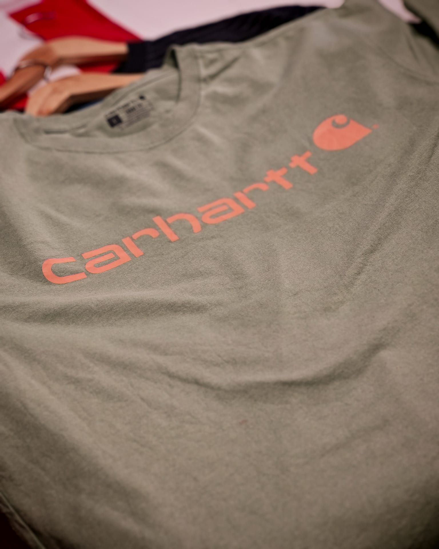 Una maglietta grigia della Carhartt con logo arancione è appoggiata su un letto.