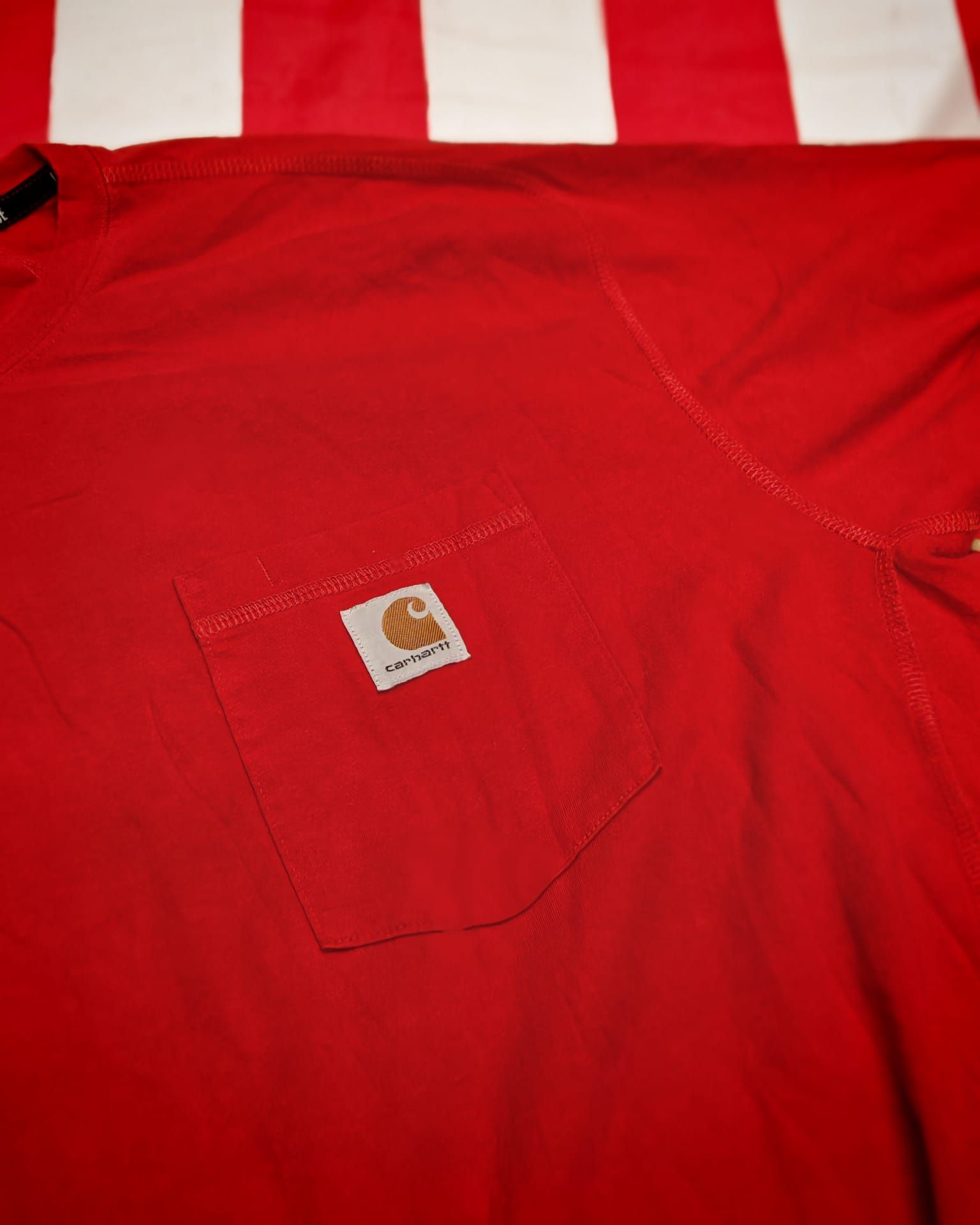Giacca Carhartt rossa con logo sulla tasca sul petto, appoggiata su una superficie a righe rosse e bianche.
