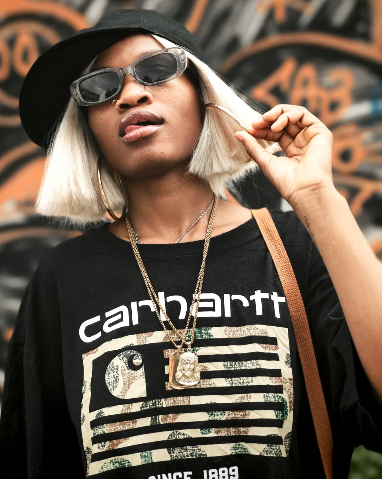 Persona con maglietta nera Carhartt e occhiali da sole, con caschetto biondo, che si tiene i capelli contro un muro ricoperto di graffiti.