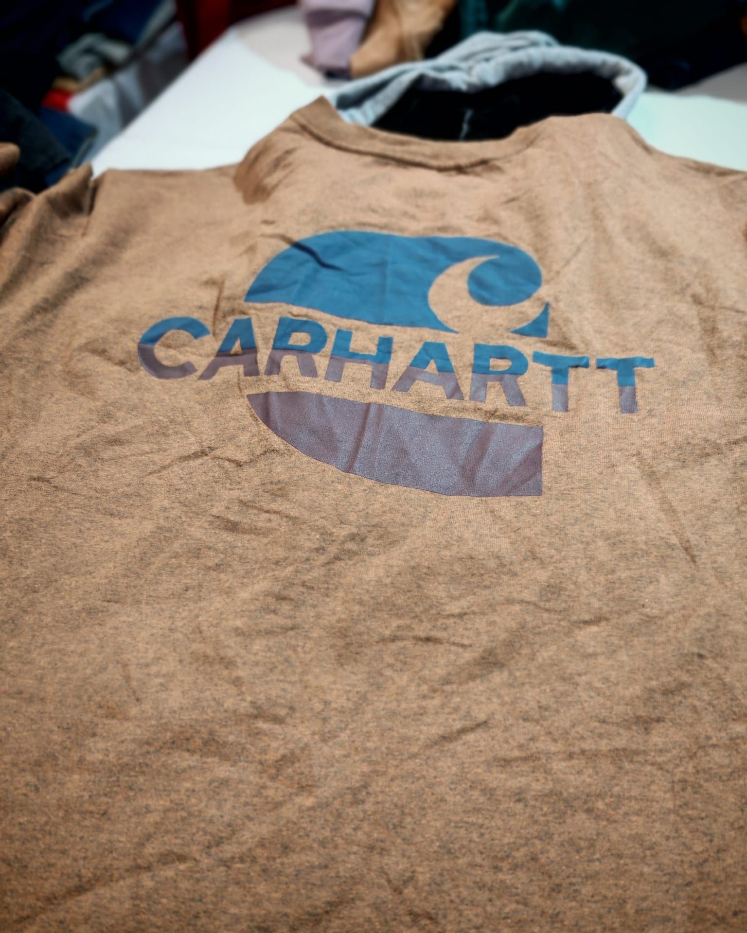 T-shirt beige Carhartt con logo blu, appoggiata su un tavolo.