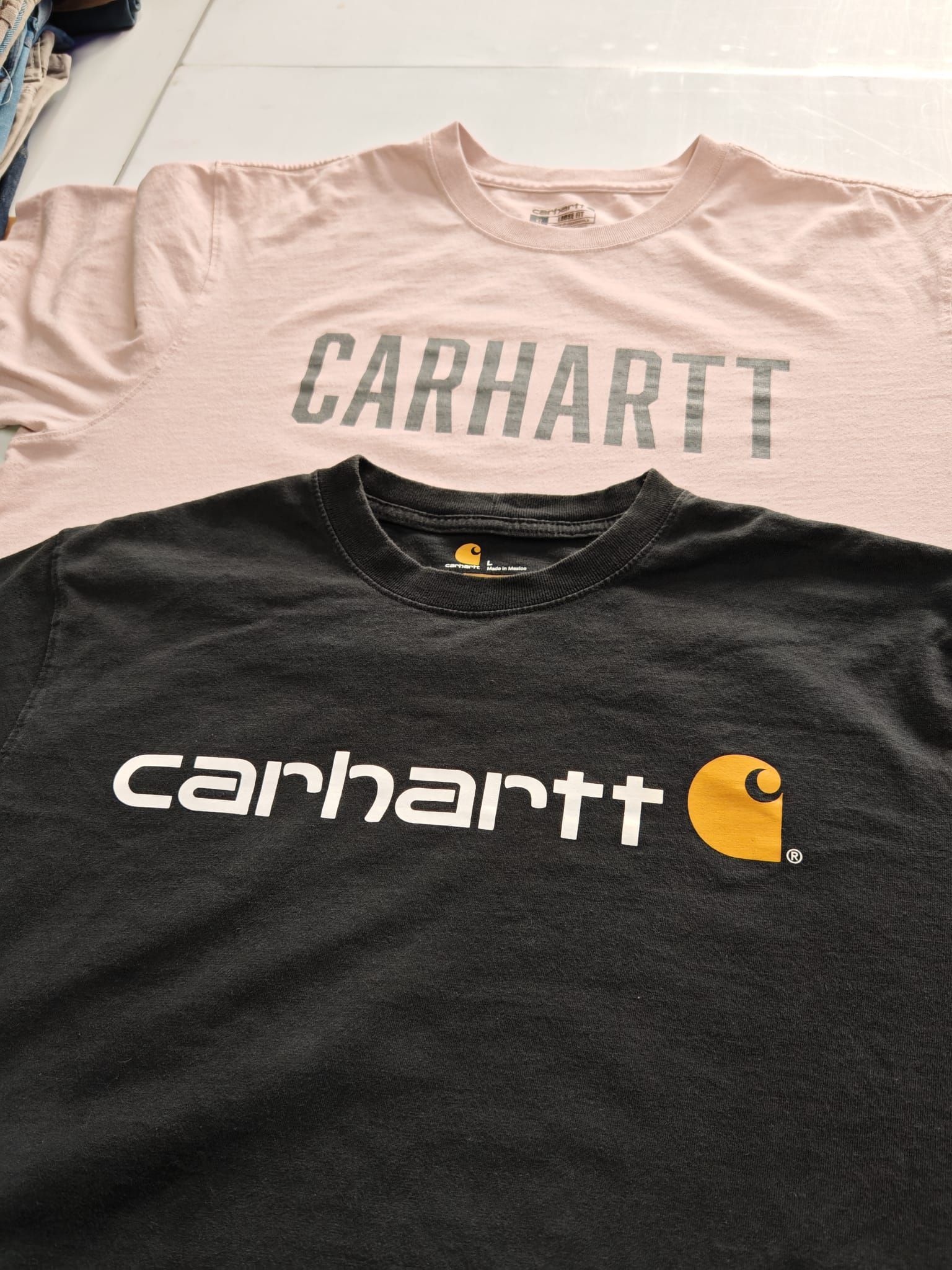 Due magliette Carhartt disposte una sull'altra: una rosa pallido con logo grigio e l'altra nera con logo bianco ed emblema arancione.