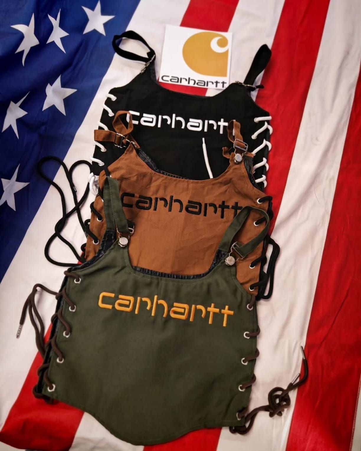 Tre grembiuli Carhartt nei colori verde oliva, marrone e beige, adagiati su uno sfondo con la bandiera americana.