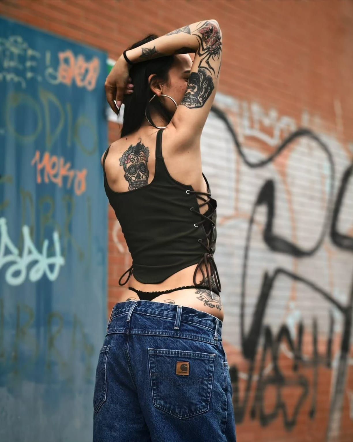 Una persona tatuata posa contro un muro di mattoni ricoperto di graffiti, indossando una maglietta nera e jeans blu.