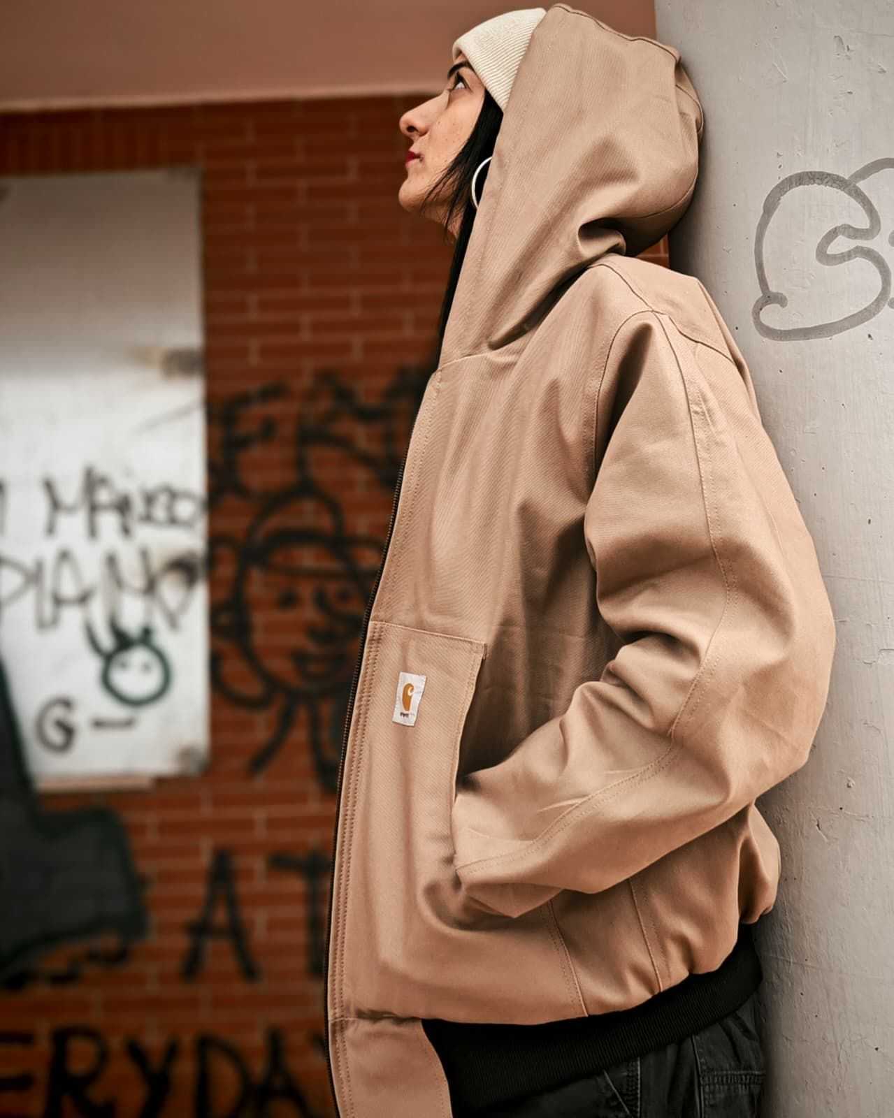 Una persona con una giacca con cappuccio color cachi è appoggiata a un muro in un contesto urbano.