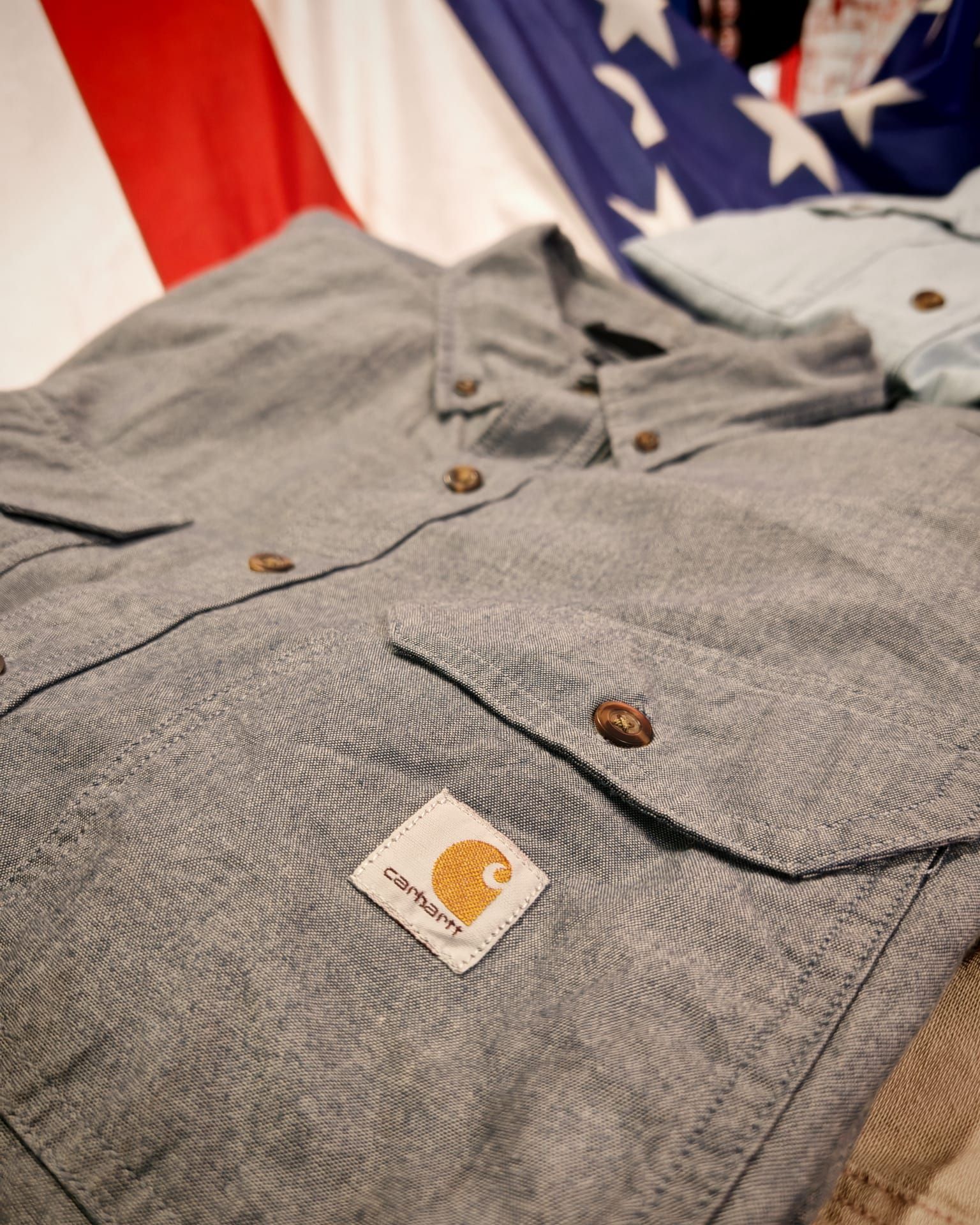 Camicia grigia abbottonata con tasche sul petto e patch con logo Carhartt, appoggiata su un tessuto con una bandiera americana sullo sfondo.