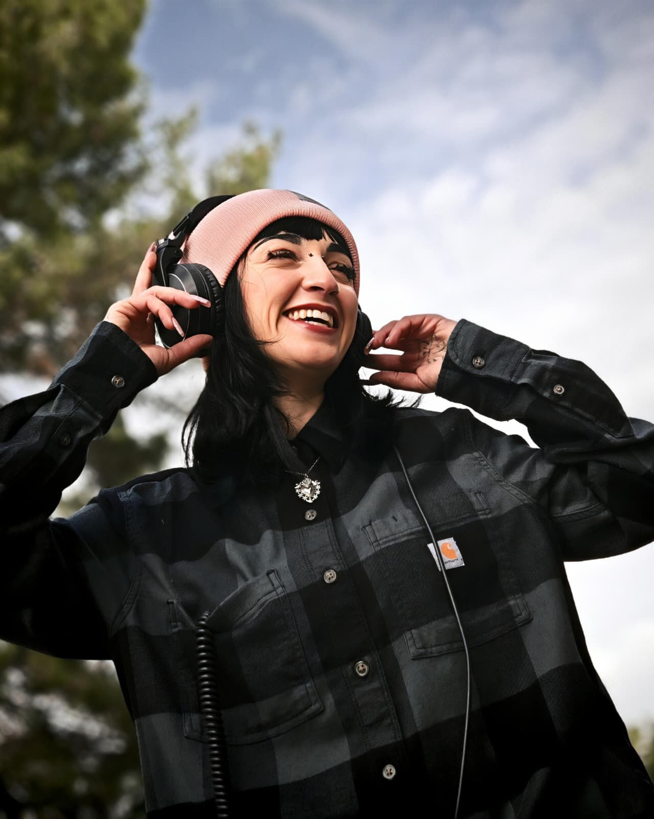 Una persona sorridente con un berretto rosa e una giacca nera si sistema le cuffie all'aperto.
