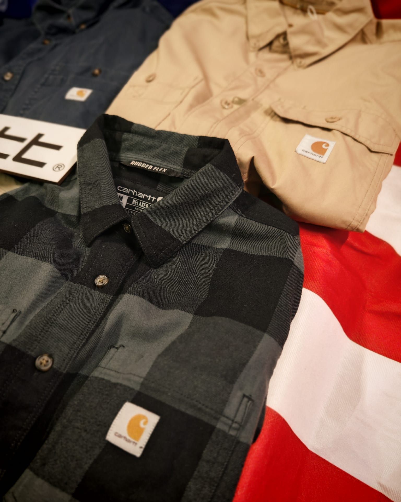 Due camicie Carhartt abbottonate, una a quadri beige e una nera, disposte su una superficie rossa e bianca
