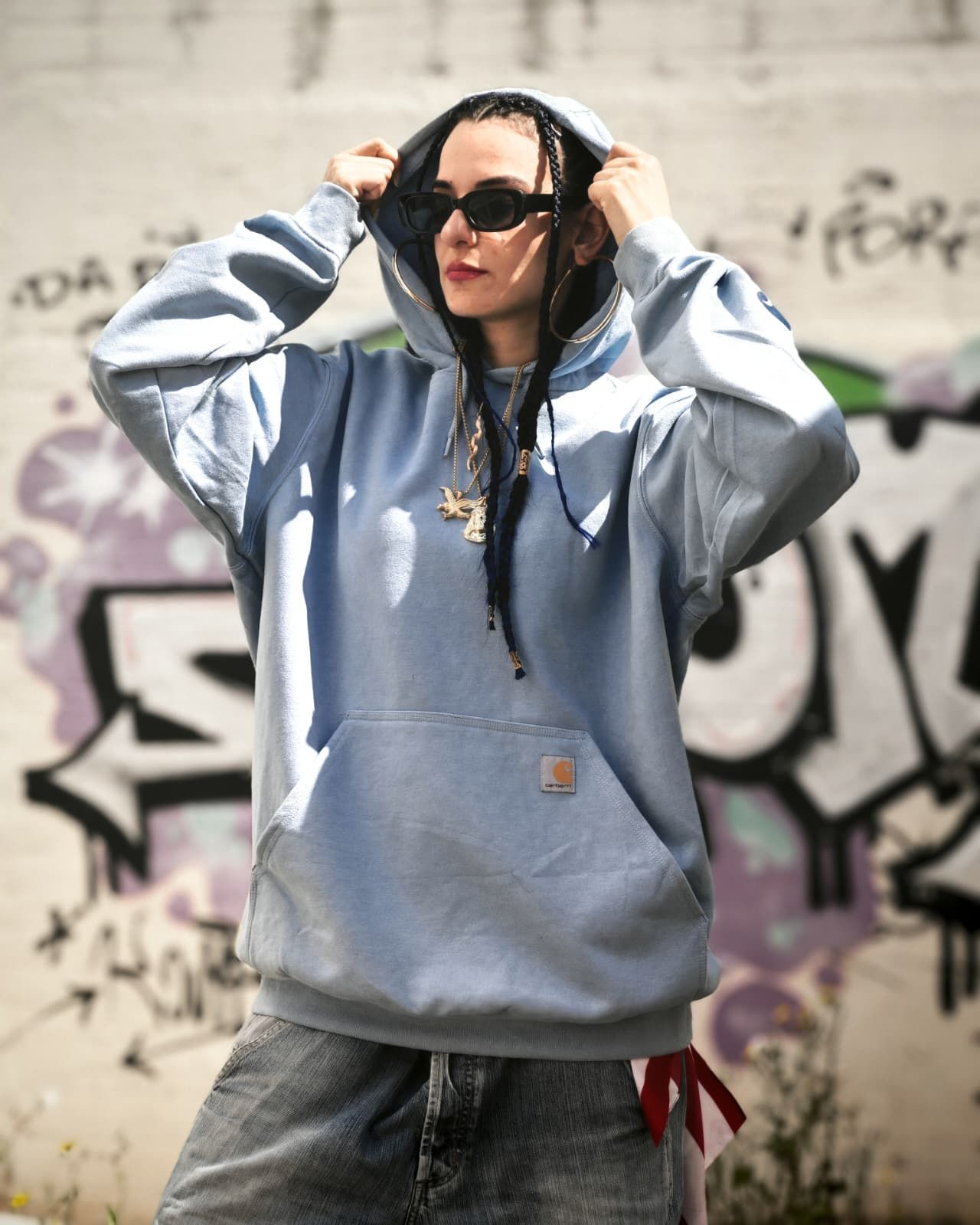 Una persona con una felpa grigia con cappuccio e occhiali da sole si sistema il cappuccio davanti a un muro ricoperto di graffiti.