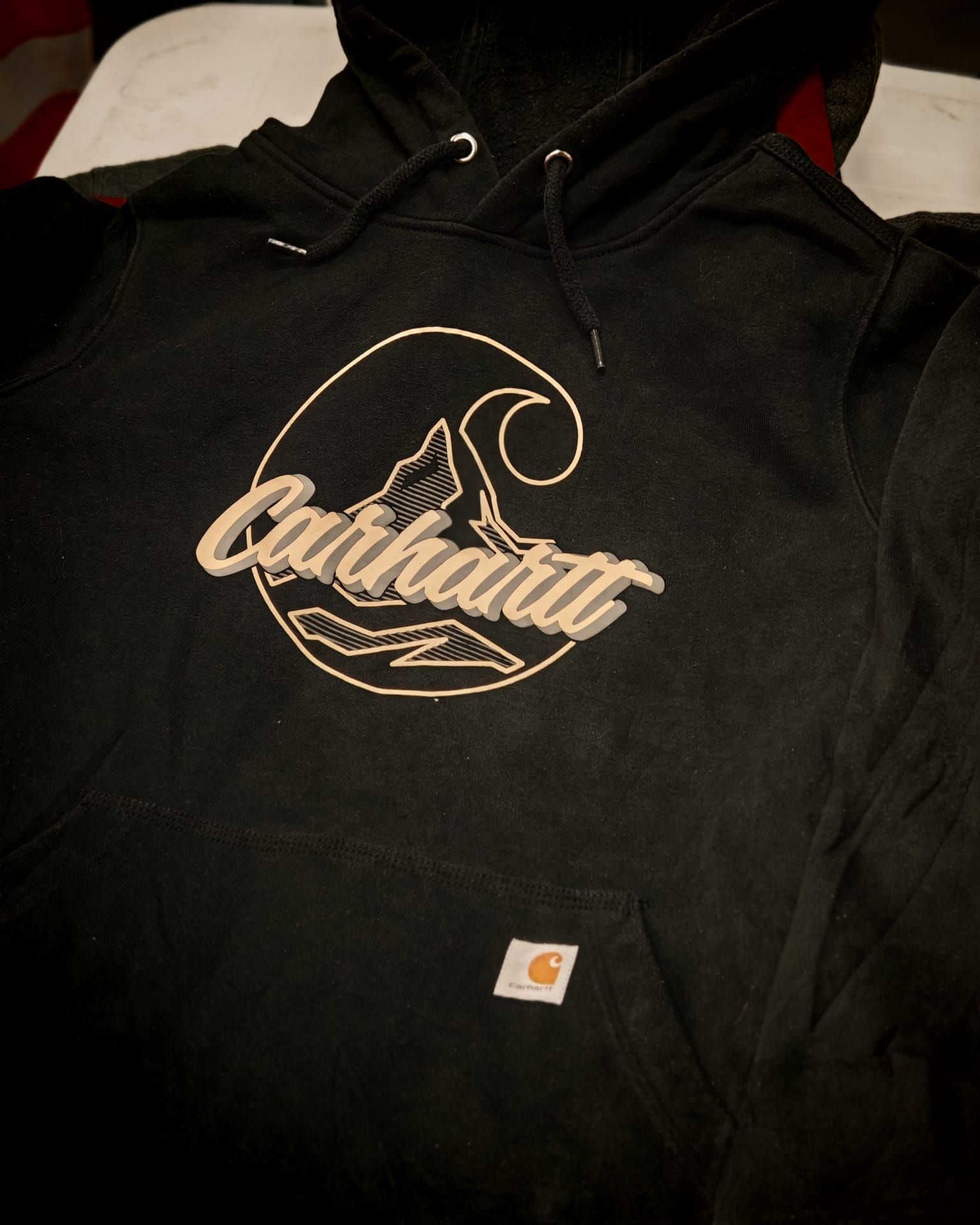 Felpa con cappuccio nera Carhartt con logo beige e piccola etichetta sul petto, distesa su un letto.