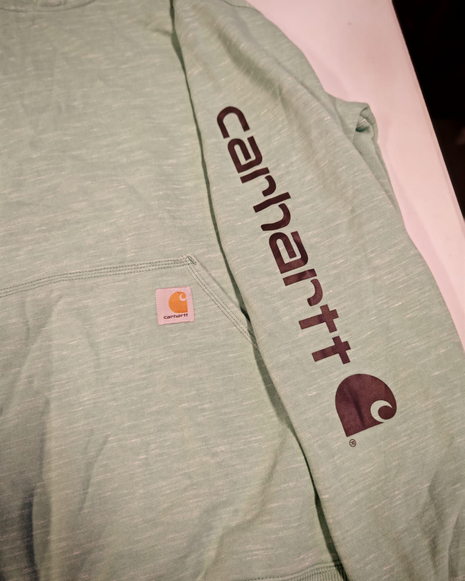 Felpa grigia Carhartt con logo nero e piccola toppa arancione, appoggiata su una superficie rosa.