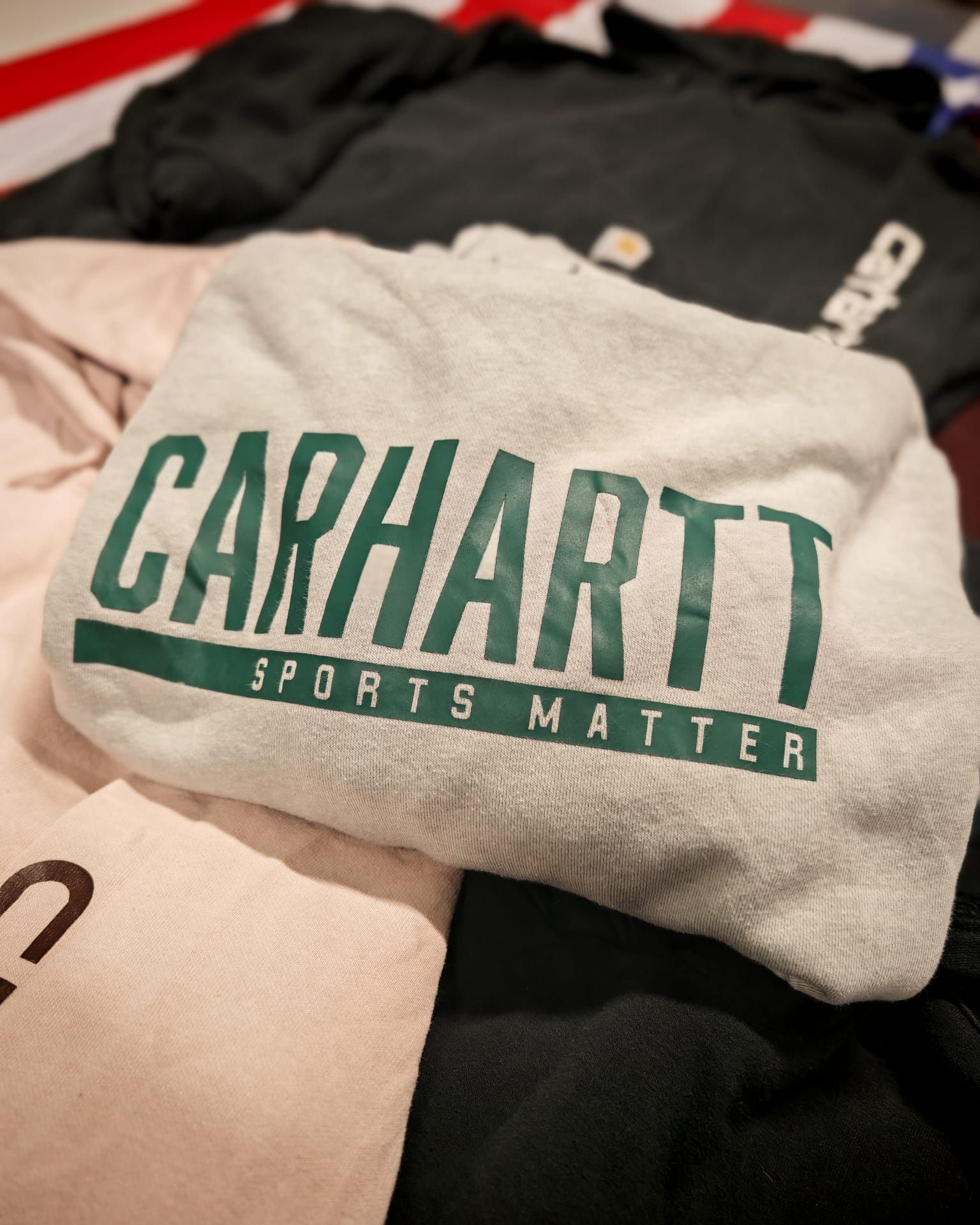 Camicia grigia Carhartt piegata con logo verde sopra una pila di indumenti.