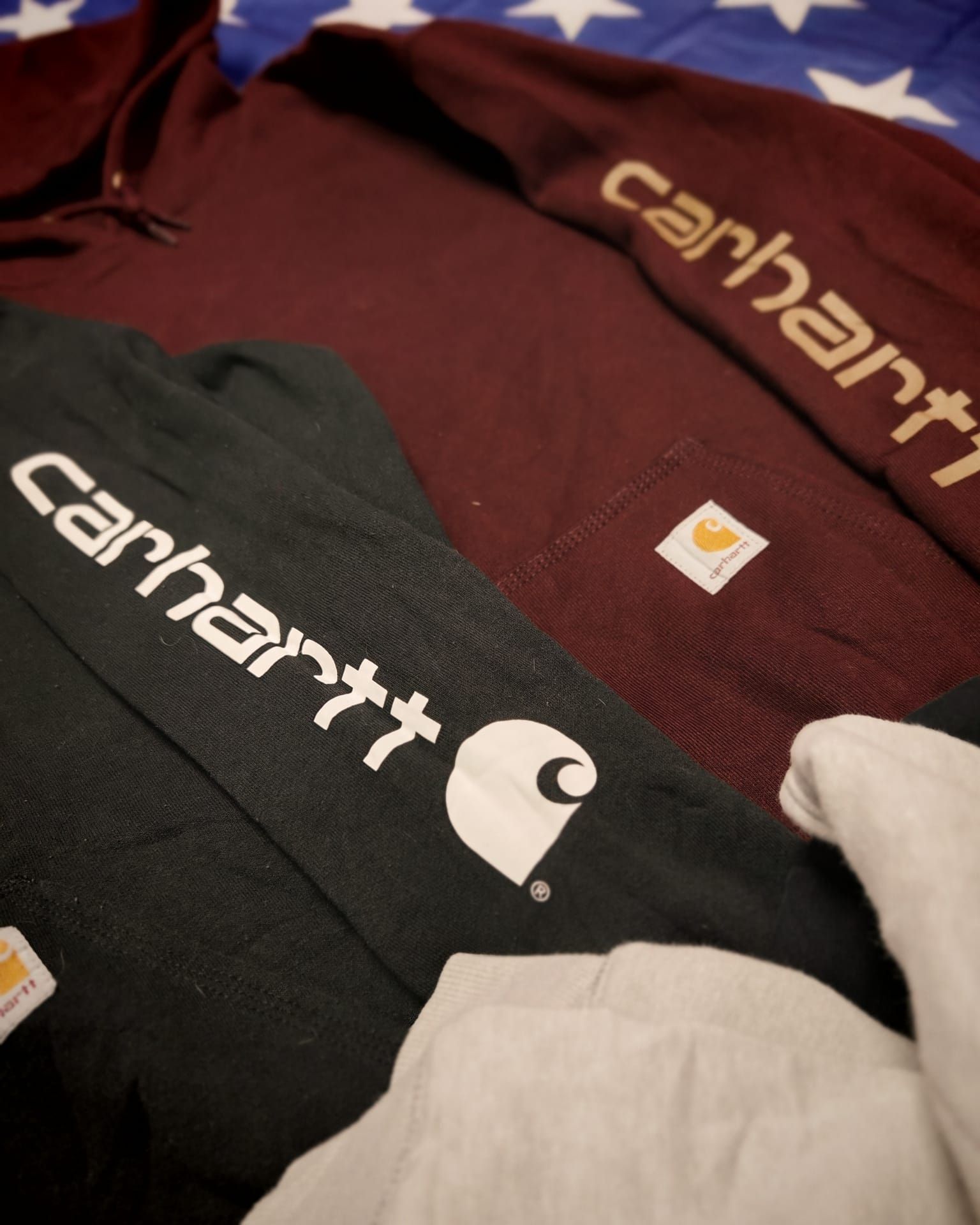 Felpe con logo Carhartt nei colori bordeaux, nero e crema disposte su uno sfondo con la bandiera americana.